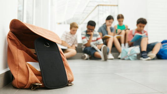 Handytasche für Schülerinnen und Schüler hilft beim Lernen. Handytasche liegt vor einem braunen Schulrucksack. Fünf Schüler lernen zusammen im Hintergrund.